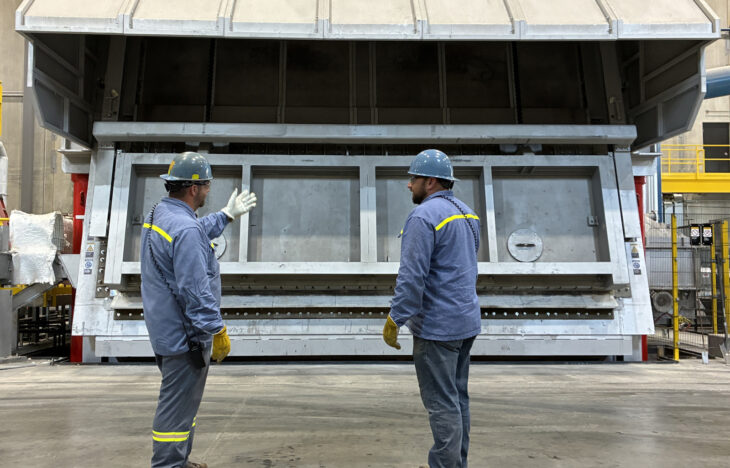 Two men stand in front of a closed aluminum furance.