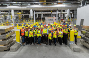 A group of people in safety gear standing in front of casting equipment with large aluminum ingot in the foreground.