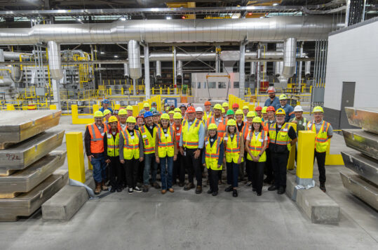A group of people in safety gear standing in front of casting equipment with large aluminum ingot in the foreground.