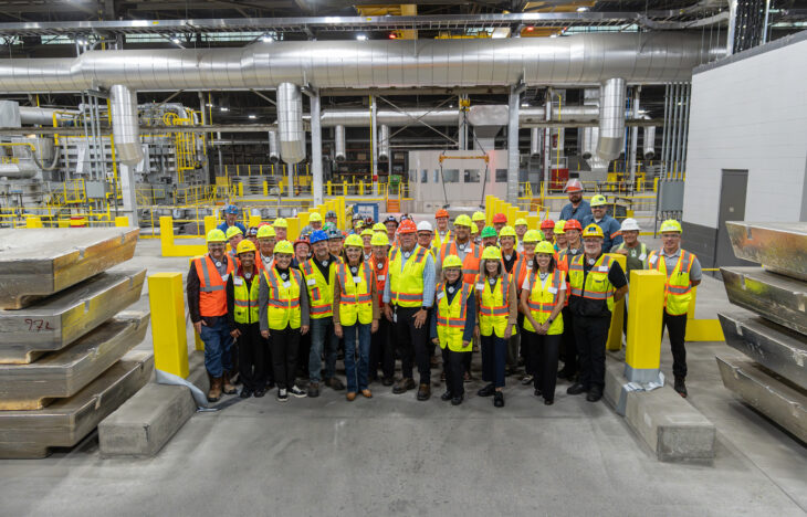 A group of people in safety gear standing in front of casting equipment with large aluminum ingot in the foreground.