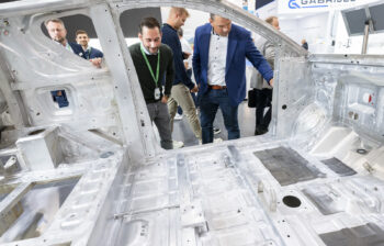 Two men in suits look into the raw aluminum spaceframe of a vehicle.