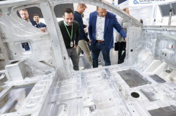 Two men in suits look into the raw aluminum spaceframe of a vehicle.