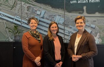 Three women stand in front of a large poster, showing an aerial view of an aluminum smelter.
