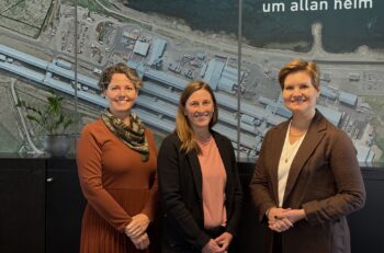 Three women stand in front of a large poster, showing an aerial view of an aluminum smelter.