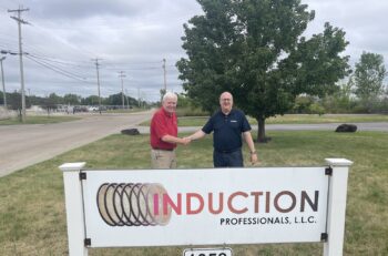 Two men shake hands, while standing by a sign that reads Induction Professionals LLC