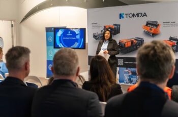 A woman stands at the front of a room talking. Behind her are images of Tomra's sorting equipment.