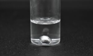 Photo of a small glass container with clear liquid and a drop of gallium metal in the bottom.