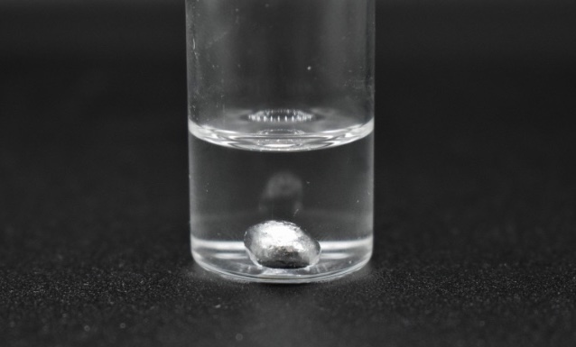 Photo of a small glass container with clear liquid and a drop of gallium metal in the bottom.