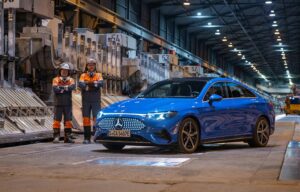 A bright blue Mercedes-Benz inside an aluminum smelter. Two ment in orange safety gear stand beside the car, smiling with crossed arms.
