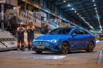 A bright blue Mercedes-Benz inside an aluminum smelter. Two ment in orange safety gear stand beside the car, smiling with crossed arms.