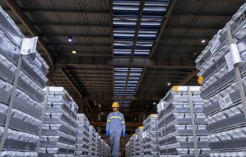 A worker in a yellow hard hat walks between stacks of aluminum ingots.