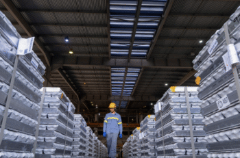 A worker in a yellow hard hat walks between stacks of aluminum ingots.