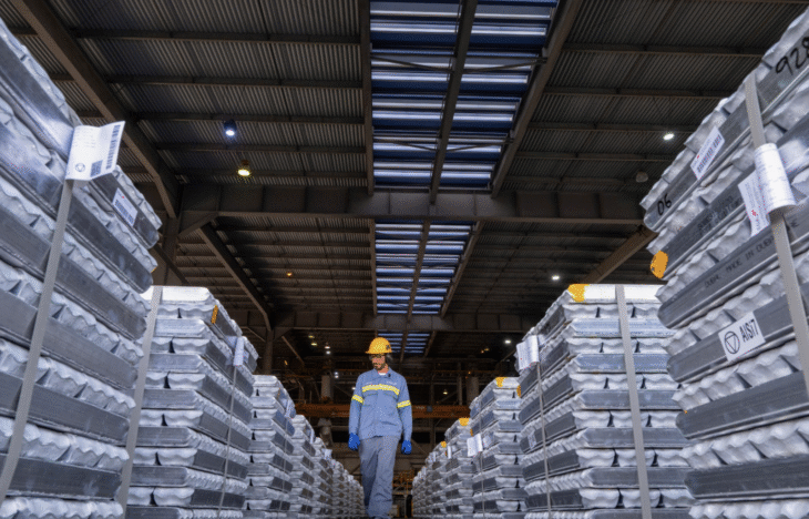 A worker in a yellow hard hat walks between stacks of aluminum ingots.