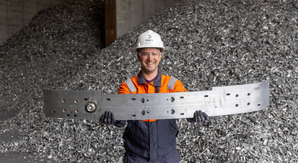 Hydro Cassopolis employee Keith Bonner holding the Cadillac OPTIQ bumper. Piles of aluminum scrap are in the background.