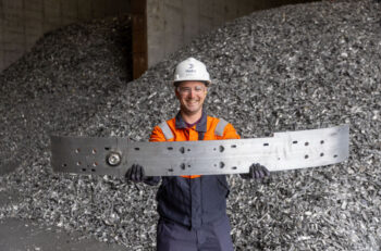 Hydro Cassopolis employee Keith Bonner holding the Cadillac OPTIQ bumper. Piles of aluminum scrap are in the background.