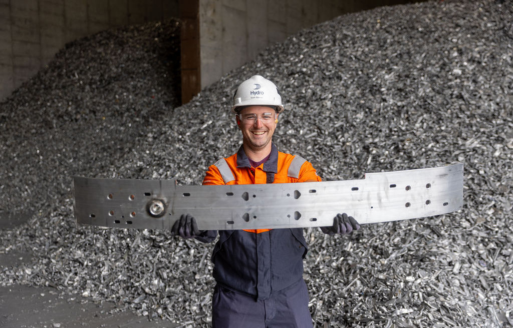 Hydro Cassopolis employee Keith Bonner holding the Cadillac OPTIQ bumper. Piles of aluminum scrap are in the background.